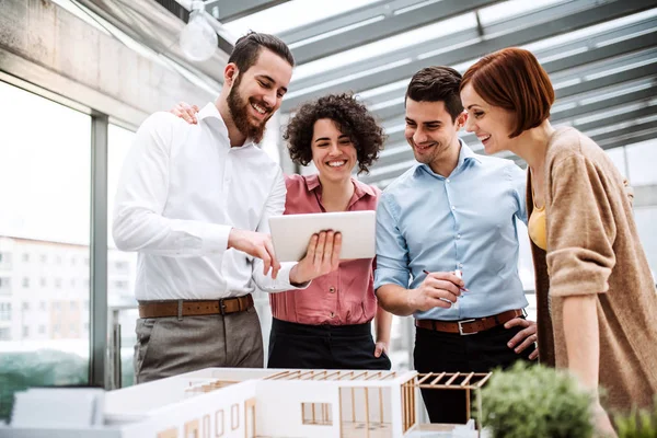 Grupo de jóvenes arquitectos con tableta y modelo de una casa de pie en la oficina, hablando . — Foto de Stock