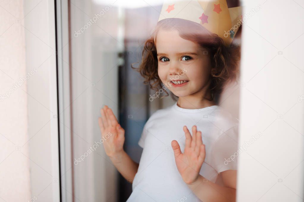 Portrait of small girl with crown at home, looking out of a window. Shot through glass.