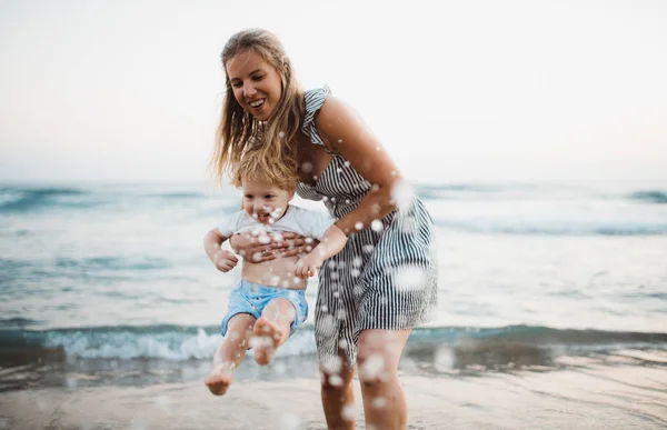 Madre joven con un niño pequeño de pie en la playa en las vacaciones de verano, divirtiéndose . —  Fotos de Stock