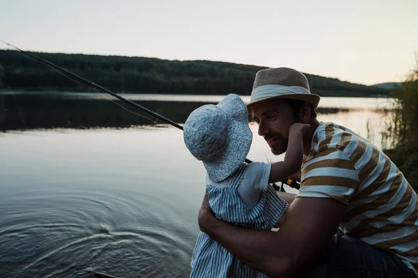 A mature father with a small toddler daughter outdoors fishing by a lake. — Stock Photo, Image
