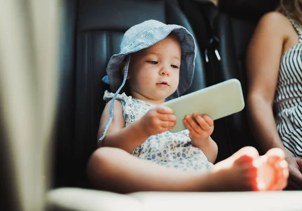 Una niña pequeña sentada en un coche, jugando con un teléfono inteligente . — Foto de Stock