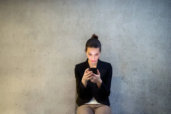 Una vista frontale della giovane donna d'affari con smartphone seduta contro il muro di cemento in ufficio . — Foto Stock