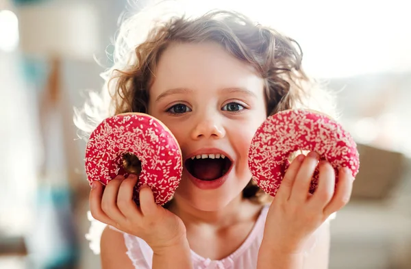 Uma menina pequena com donuts em casa, olhando para a câmera . — Fotografia de Stock