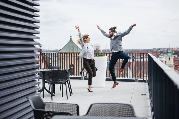 Two young business people jumping on terrace outside office, expressing excitement.