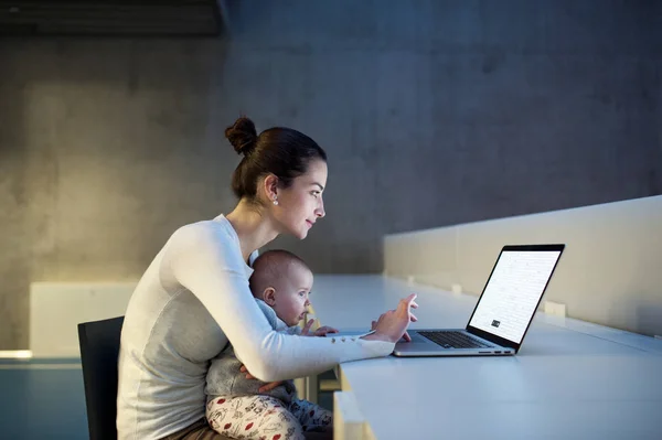 Jovem estudante com um bebê sentado na mesa em um quarto em uma biblioteca ou escritório, usando laptop . — Fotografia de Stock