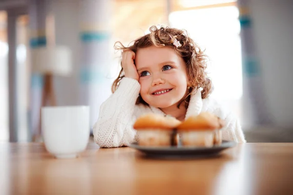 Um retrato de menina pequena com muffins sentados à mesa em casa . — Fotografia de Stock