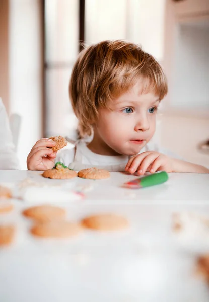 Un niño pequeño sentado a la mesa, decorando y comiendo pasteles en casa . —  Fotos de Stock