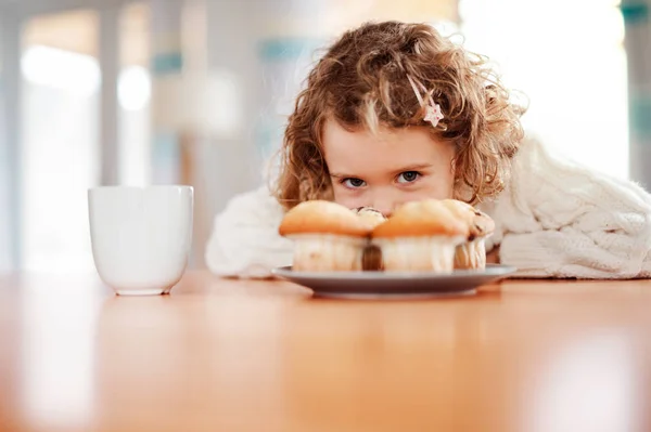 Un retrato de una niña pequeña con magdalenas sentada a la mesa en casa . — Foto de Stock