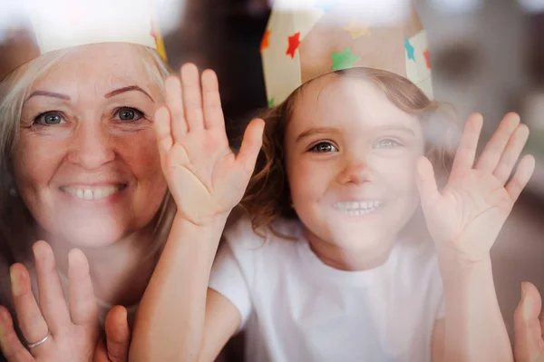 A portrait of small girl with grandmother having fun at home. Shot through glass. — Stock Photo, Image