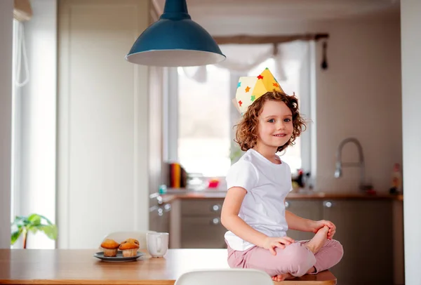 Una niña pequeña con una corona de papel en casa, mirando a la cámara . — Foto de Stock