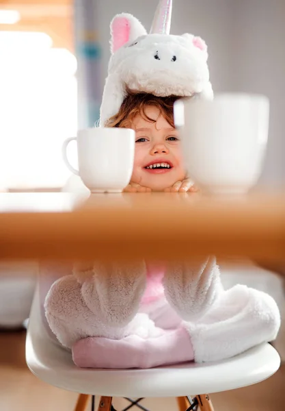 Een portret van een klein meisje in unicorn masker aan de tafel thuis. — Stockfoto