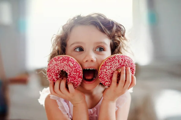 Una bambina con le ciambelle a casa, guardando la macchina fotografica . — Foto Stock