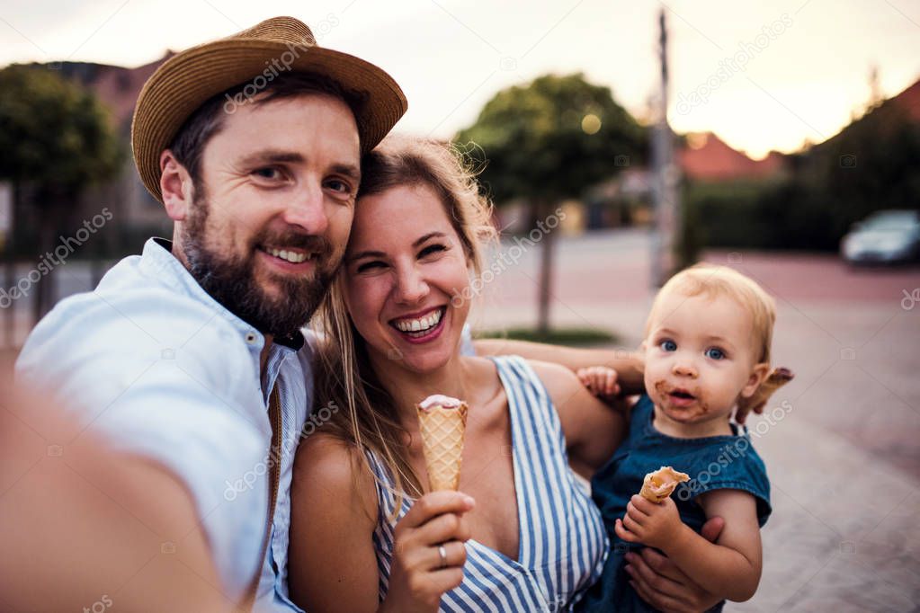 Parents and small toddler girl with ice cream outdoors in summer, taking selfie.
