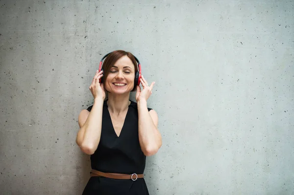 Young business woman with headphones standing against concrete wall in office. — Stock Photo, Image