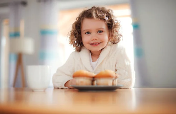 Ett porträtt av liten flicka med muffins sitter vid bordet hemma. — Stockfoto