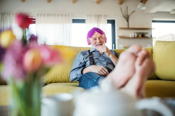 En senior man med hörlurar och ögonmask sitter på soffan inomhus hemma. — Stockfoto