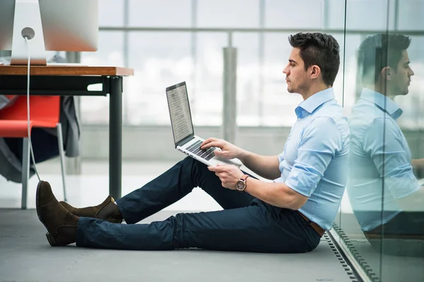 Um jovem homem de negócios com laptop sentado no chão no escritório, trabalhando . — Fotografia de Stock