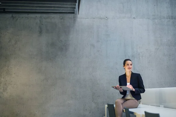 Młoda kobieta biznesu z tabletem siedzi na biurku pod betonową ścianą w biurze. — Zdjęcie stockowe