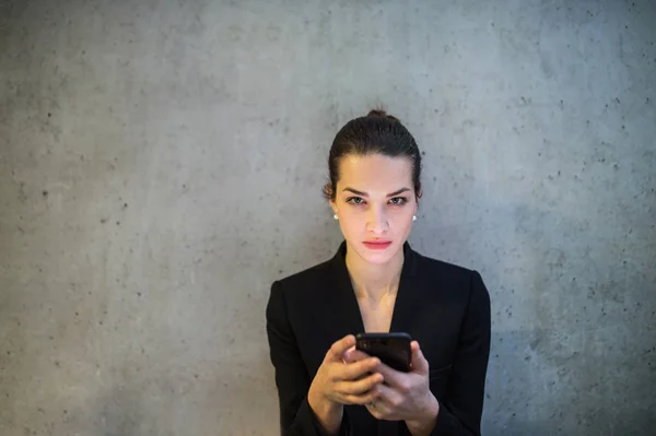 Una vista frontale di giovane donna d'affari con smartphone in piedi contro muro di cemento in ufficio . — Foto Stock