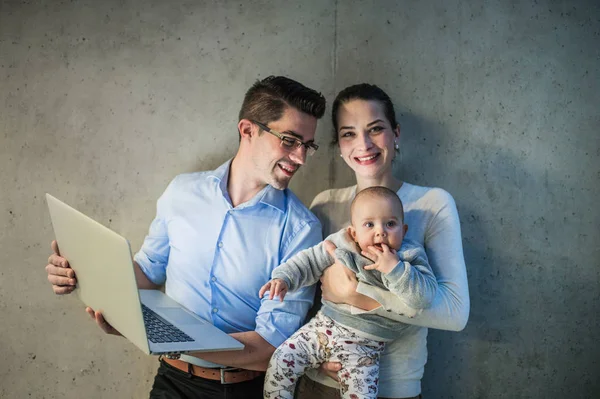 Un joven hombre de negocios con esposa e hija bebé de pie en la oficina, utilizando el ordenador portátil . —  Fotos de Stock