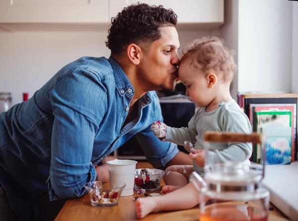 A father and a small toddler son eating fruit and yoghurt indoors at home. — Stock Photo, Image