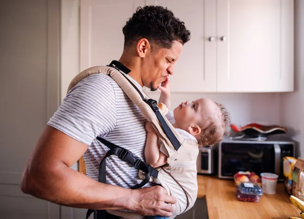 Un padre con un pequeño hijo en el portador en la cocina en el interior de casa . —  Fotos de Stock