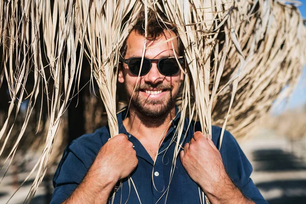 Un uomo maturo con gli occhiali da sole in piedi all'aperto sulla spiaggia, ombrello di paglia sulla testa . — Foto Stock