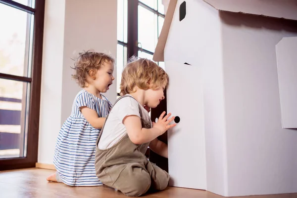 Due bambini piccoli che giocano con una casa di carta di cartone all'interno di casa . — Foto Stock