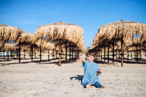 Malá holčička seděla na písku mezi slámově slunečníky venku na pláži. — Stock fotografie