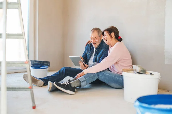 Pareja de ancianos pintando paredes en un nuevo hogar, usando una tableta. Concepto de reubicación . — Foto de Stock