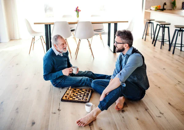 Un fils hipster adulte et père aîné assis sur le sol à la maison, jouant aux échecs . — Photo