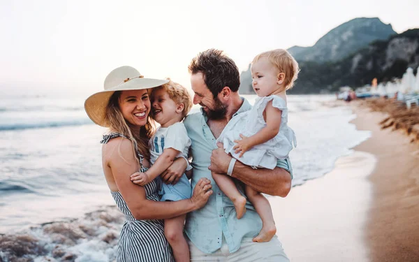 Una familia joven con dos niños pequeños de pie en la playa en las vacaciones de verano . — Foto de Stock