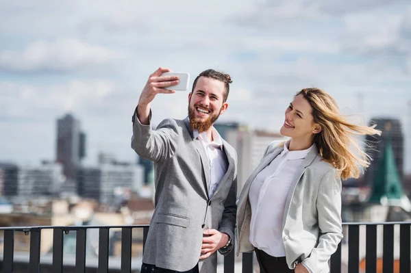 スマートフォンを持つ2人の若いビジネスマンがテラスに立って、自分撮りを撮ります. — ストック写真