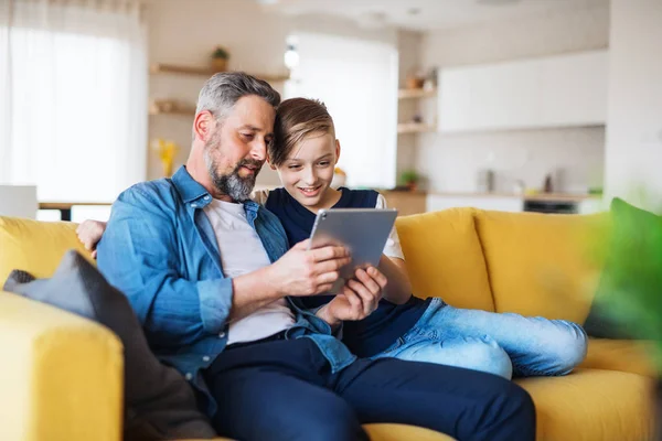 Mature father with small son sitting on sofa indoors, using tablet. — Stock Photo, Image