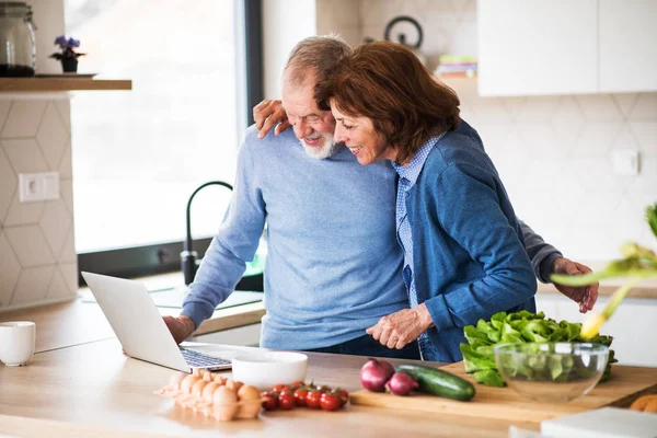 Ett porträtt av Senior par med laptop inomhus hemma, Matlagning. — Stockfoto