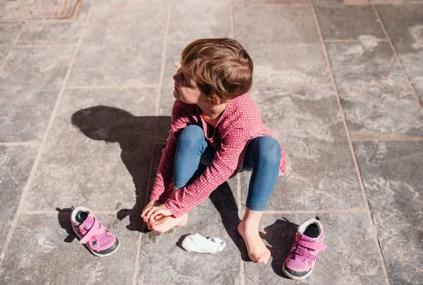 Вид сверху на маленькую девочку, сидящую на улице на тротуаре, снимающую обувь . — стоковое фото