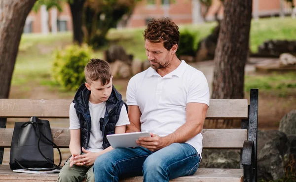 Padre con hijo sentado en el banco al aire libre en la ciudad, usando la tableta . —  Fotos de Stock