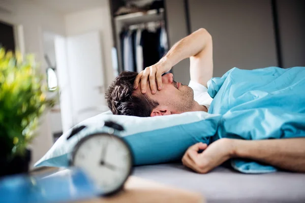 Young tired man with alarm clock in bed at home, waking up in morning. — Stock Photo, Image