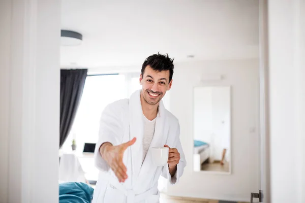 Mladý muž s kávou nabízí ruku pro pozdrav v v ložnici, ranní rutina. — Stock fotografie