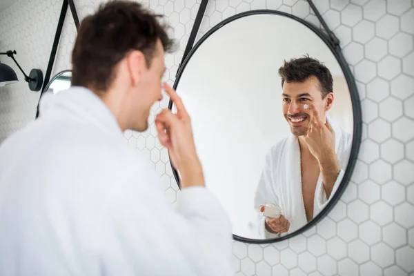 Jeune homme mettre de la crème sur le visage dans la salle de bain le matin, routine quotidienne . — Photo