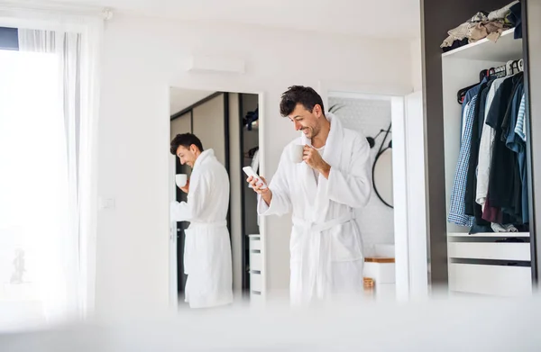 Молодий чоловік з кавою і смартфоном в спальні, ранкова рутина . — стокове фото