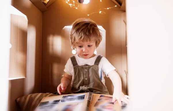 Bambino che gioca al chiuso in una casa di cartone a casa, guardando l'album fotografico . — Foto Stock