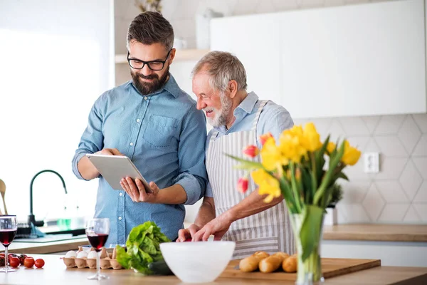 Hijo hipster adulto y padre mayor en el interior de la cocina en casa, utilizando tableta . —  Fotos de Stock