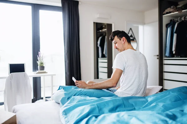 Jovem com smartphone na cama em casa, mensagens de texto . — Fotografia de Stock