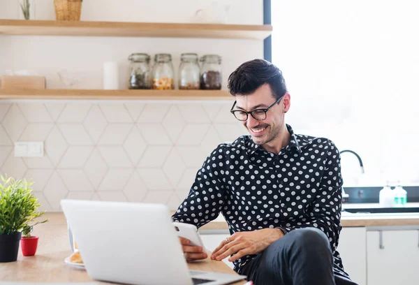 Young man with laptop and smartphone sitting in kitchen, a home office concept. — Stock Photo, Image