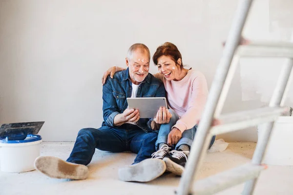 Pareja de ancianos pintando paredes en un nuevo hogar, usando una tableta. Concepto de reubicación . — Foto de Stock