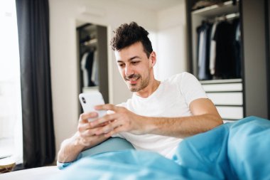 Evde yatakta akıllı telefon ile genç adam, metin mesajlaşma.