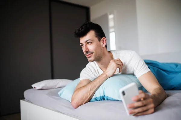 Jovem com smartphone na cama em casa, mensagens de texto . — Fotografia de Stock