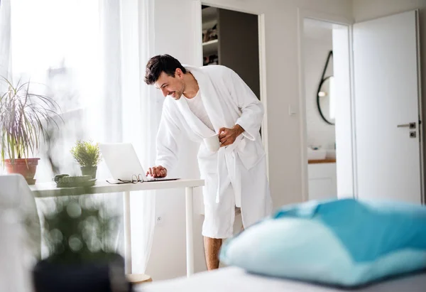 Young man with laptop and coffee in the bedroom in the morning. — Stock Photo, Image