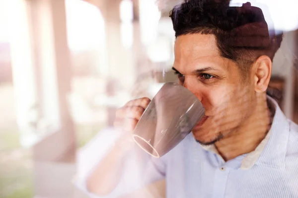 Um homem dentro de casa, à janela em casa, a beber café. Tiro através de vidro . — Fotografia de Stock